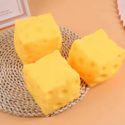 Three yellow cheese-shaped foam cubes with realistic holes, arranged on a woven round mat against a peach-colored background.