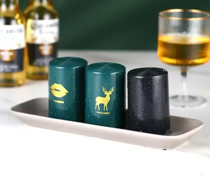 Three cylindrical condiment or sauce containers with a water-droplet finish sit on a long rectangular dish. Two containers are dark green with gold printed graphics (lips and deer), and one is dark gray/black.