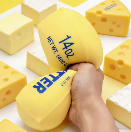 A hand squeezing a yellow novelty cheese-shaped squishy toy, with other cheese-shaped squishies scattered around on a white surface.