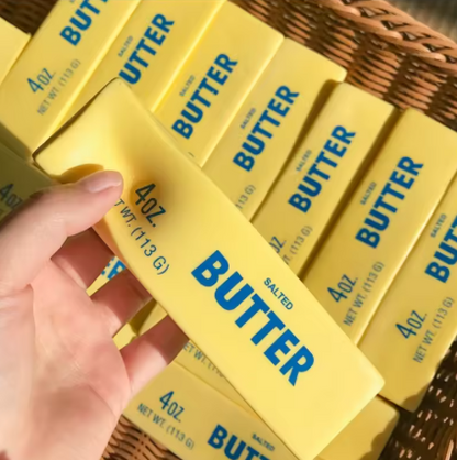 A hand holding a yellow 4 oz package of salted butter, with multiple matching butter packages arranged in a woven basket background.