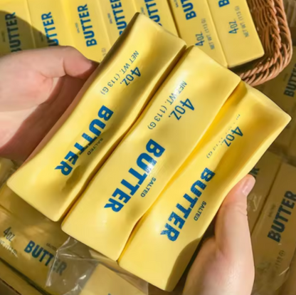 Hands holding several yellow butter sticks with blue 'BUTTER' text, with more butter and a woven basket in the background.