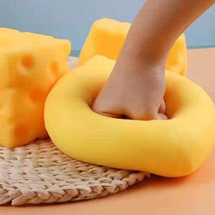 A yellow donut-shaped foam cushion with a hand pressing into it, shown alongside two yellow cheese-shaped cushions on a woven mat against a soft blue and peach background.