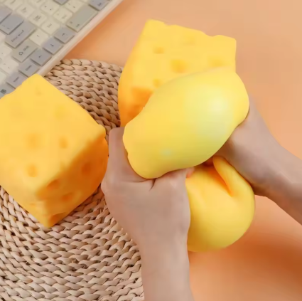Hands squeezing a yellow squishy toy in front of two cheese-shaped stress toys on a woven placemat, with part of a keyboard visible in the background.