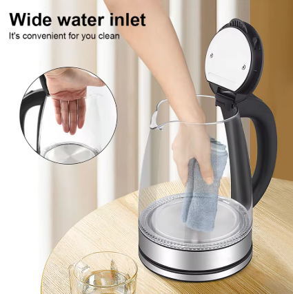 A glass electric kettle with a black handle and base sits on a wooden table in a kitchen setting. A person's arm is shown wiping the inside of the kettle with a cloth, and a circular inset highlights the wide water inlet.
