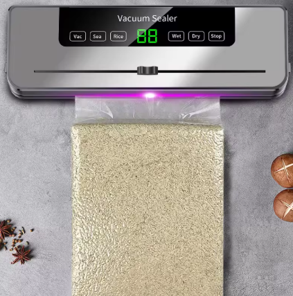 A countertop vacuum sealer machine with a metallic silver body and black control panel, shown sealing a clear plastic bag containing a light beige textured item. The background is a gray stone-like surface with a few decorative spices on the left and two mushrooms on the right.