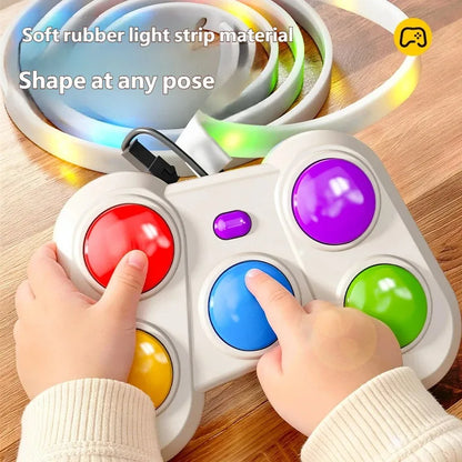 A child's hands press colorful round buttons on a white handheld toy controller. A coiled multicolor light strip is shown behind it on a wooden surface, with text reading 'Soft rubber light strip material' and 'Shape at any pose.