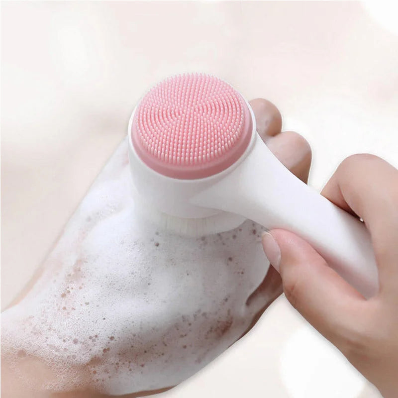 A handheld facial cleansing brush with a white handle and pink silicone bristles being used on a foamy hand, shown against a soft light background.
