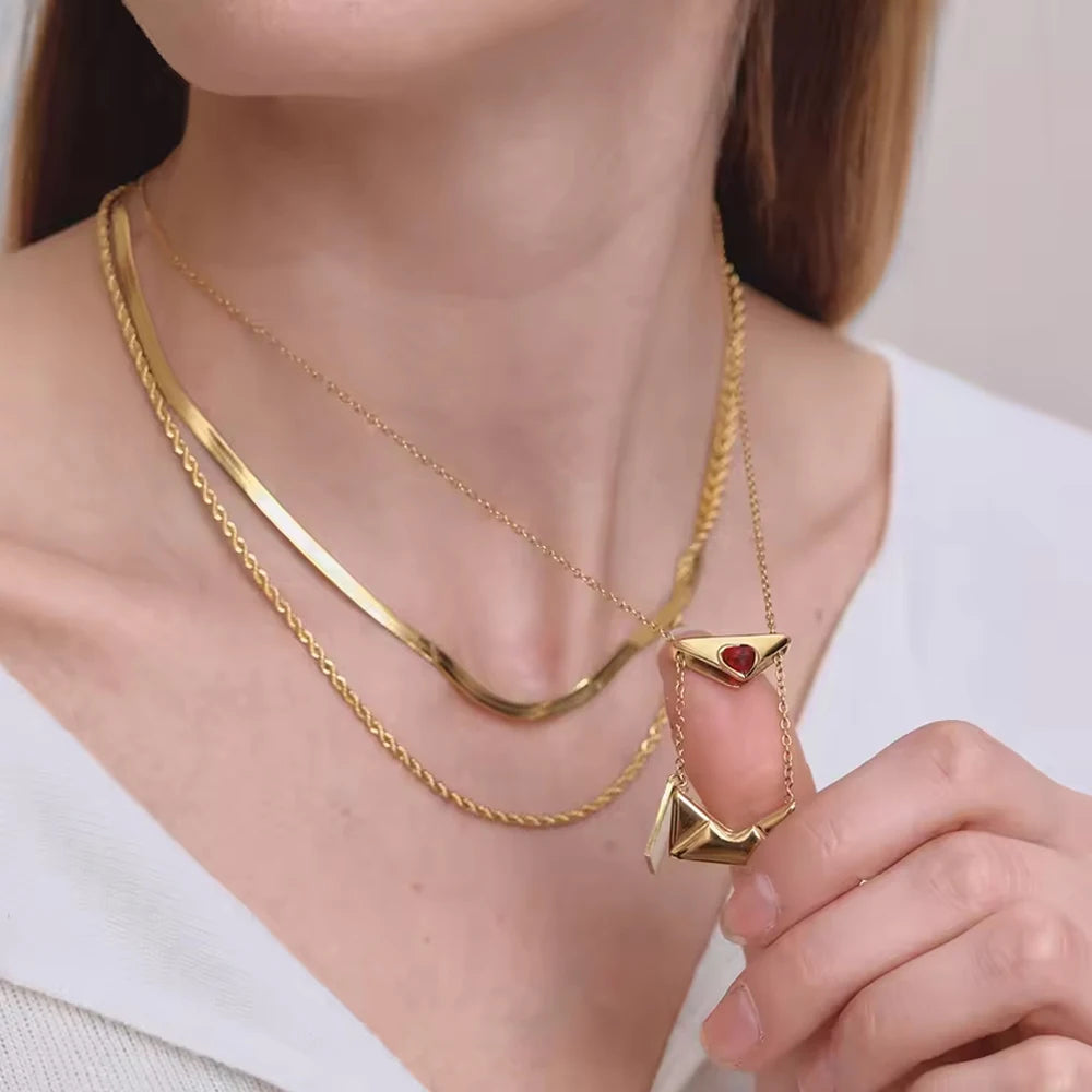 Gold necklace with a red heart pendant held by a hand, worn by a person.