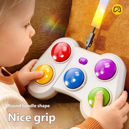 A child holds a white toy game controller with colorful round buttons while sitting on a brown couch. The controller has red, yellow, blue, purple, and green buttons, and a small light bar attached at the top glows yellow.