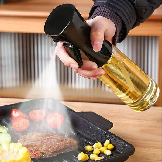 A hand holds a glass oil sprayer bottle filled with oil above a grill pan with food, spraying oil into the air. The scene is set on a wooden table with a blurred background.