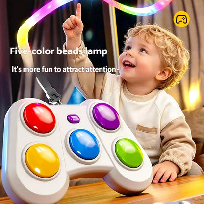 A child sitting at a table with a large white toy game controller featuring five colorful round buttons. The scene includes glowing multicolor light beads overhead and text that reads 'Five-color beads lamp' and 'It's more fun to attract attention.