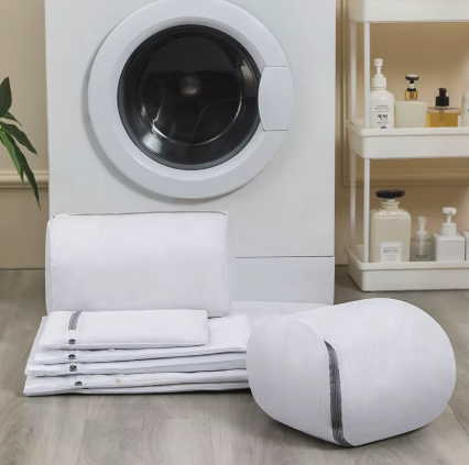 A white fabric laundry accessory set arranged on a light wood floor in front of a white front-loading washing machine, with a matching white cylindrical container or bag placed beside folded towels and a pillowcase.