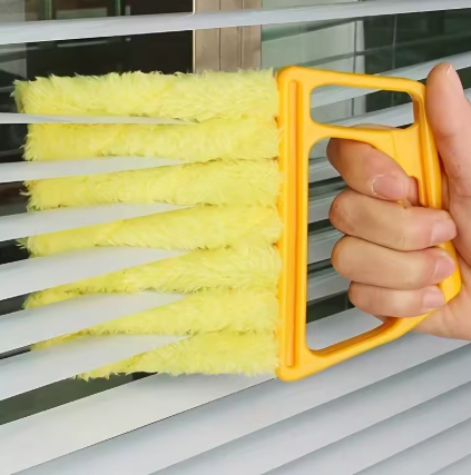 A hand holding a yellow window blind cleaning brush with yellow feather-like bristles against white window blinds.