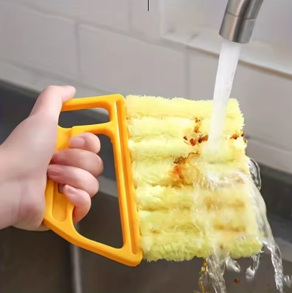 A hand holding a yellow sponge brush with a handle under running water from a faucet in a kitchen sink.